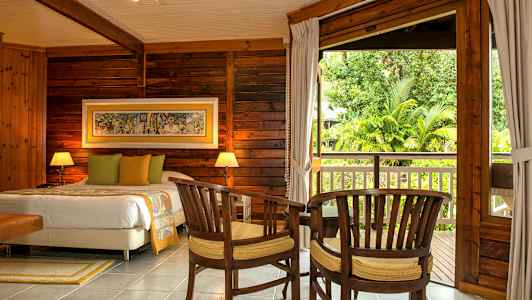 Et trepanelt soverom med en dobbeltseng, to stoler og et bord foran en stor åpning mot en balkong med hageutsikt. (AI-generert tekst)