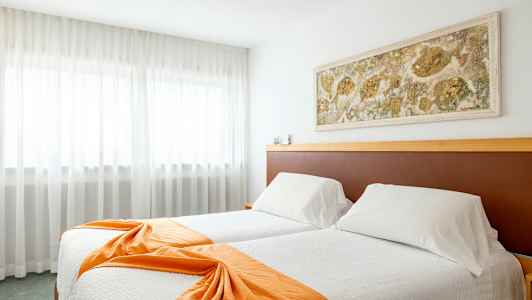 Et hotellrom med to senger kledd i hvite sengetøy og oransje dekorative sjal, en veggdekorasjon over sengene og gardiner foran et stort vindu. (AI-generert tekst)