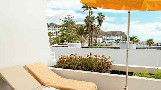 Solstol med håndkle under parasoll på en balkong med utsikt mot palmer og bygninger. (AI-generert tekst)