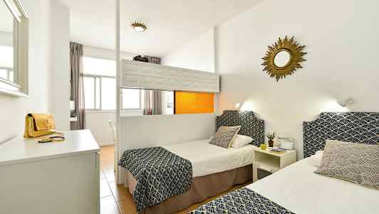 Et hotellrom med to enkeltsenger dekorert med mønstrede overkast og puter, hvite vegger og et speil på veggen. (AI-generert tekst)