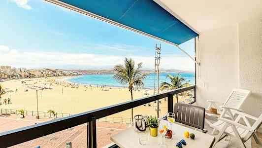 Utsikt fra en balkong mot en sandstrand og hav, med bord og stoler i forgrunnen. (AI-generert tekst)