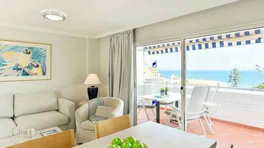 Interiør av en stue med sofa, lenestol og bord, og utsikt over havet gjennom glassdører til en terrasse. (AI-generert tekst)