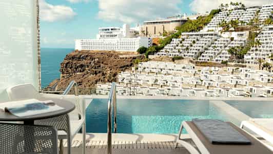 Utsikt fra en uteplass med basseng mot et fjell med bygninger. (AI-generert tekst)