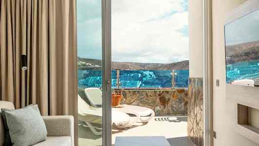 En utsikt fra et rom mot en terrasse med solsenger og glassrekkverk, landskap og himmel i bakgrunnen. (AI-generert tekst)