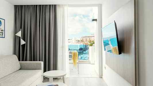 En stue med sofa, bord, TV og en åpen balkongdør som viser utsikt over bygninger og hav. (AI-generert tekst)
