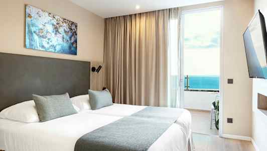 Et hotellrom med en dobbeltseng, beige gardiner og havutsikt gjennom en glassdør. (AI-generert tekst)