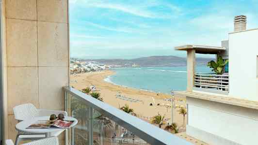 En balkong med bord og stoler gir utsikt over en sandstrand og havet. (AI-generert tekst)