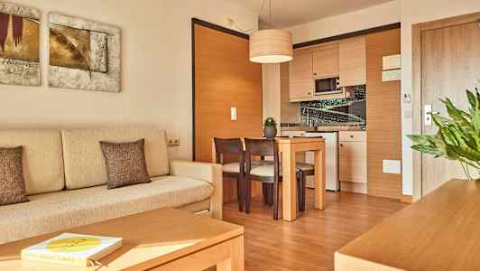 Bilde av et moderne hotellrom med en lys sofa, et trebord med stoler og en hjørnekjøkken. (AI-generert tekst)