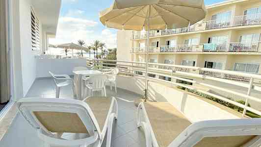 Balkong med solsenger under en parasoll og utsikt mot bygninger og palmer. (AI-generert tekst)