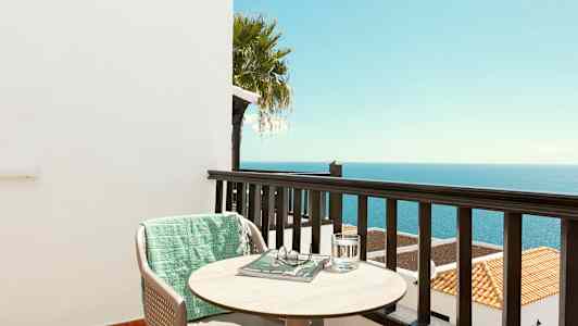 Utsikt fra en balkong mot havet med en stol, et bord og et grønt teppe. (AI-generert tekst)