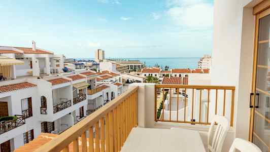 Utsikt fra en balkong med et bord og stoler over hvite bygninger med røde tak og havet i bakgrunnen. (AI-generert tekst)