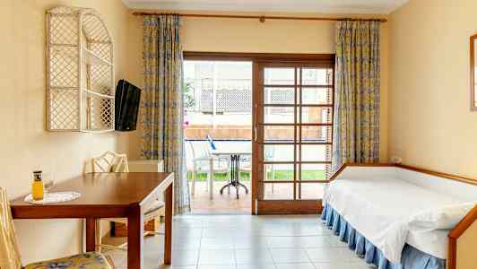 Et hotellrom med en enkeltseng, bord og stoler, samt utsikt mot en terrasse med møbler. (AI-generert tekst)