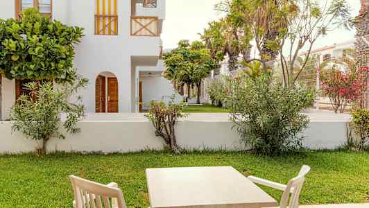 Utemøbler med bord og stoler foran en grønn gressplen og hvite bygninger. (AI-generert tekst)