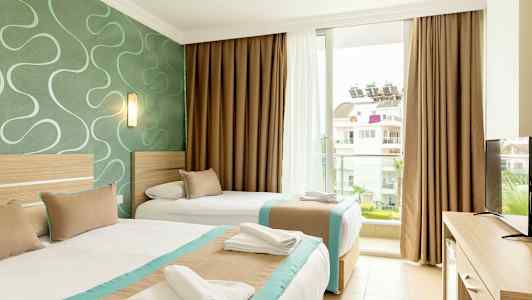 Et hotellrom med to senger. Sengene er oppredde med hvitt sengetøy og har beige og turkise sengetepper. Rommet har en grønn vegg med et mønster og tilbyr utsikt mot en bygning utenfor vinduet. (AI-generert tekst)