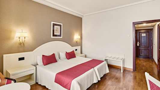 Et hotellrom med en dobbeltseng som har røde detaljer, vegglamper og en dør i bakgrunnen. (AI-generert tekst)