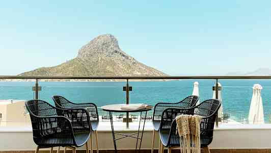 Fire stoler og et bord på en terrasse med utsikt over havet og en fjelltopp. (AI-generert tekst)