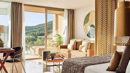 Et hotellrom med en seng, sittegruppe og utsikt over grønn natur gjennom glassdører. (AI-generert tekst)