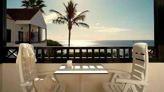 Utsikt fra en terrasse med bord og stoler mot havet, med en palme og en bygning med rødt tak i bakgrunnen. (AI-generert tekst)