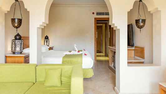 Et hotellrom med en seng dekorert med et formet håndkle og blomster, en grønn sofa i forgrunnen og utsmykkede lamper. (AI-generert tekst)