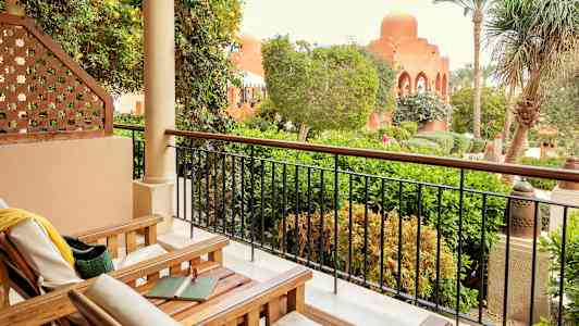 En terrasse med trestoler og utsikt over en frodig hage og bygninger med bueformede vinduer. (AI-generert tekst)