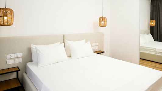 Et moderne hotellrom med en stor seng oppredet med hvite laken. Hengende lamper i rottingstil flankerer sengen. (AI-generert tekst)