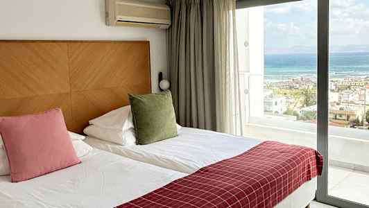 Et hotellrom med to enkeltsenger, en stor glassdør som viser utsikt over havet og fasiliteter som klimaanlegg. (AI-generert tekst)