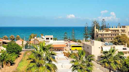 Utsikt over et boligområde med palmer og bygninger, og havet synlig i bakgrunnen under en klar blå himmel. (AI-generert tekst)