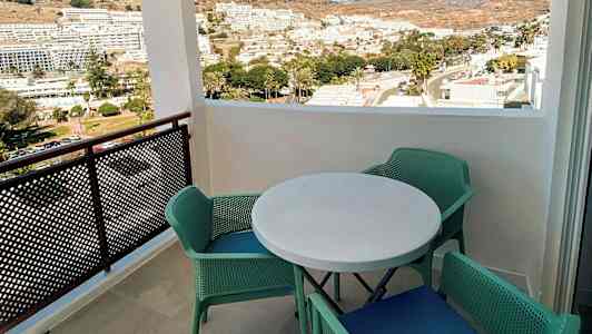 Utsikt fra en balkong med et rundt bord og fire grønne stoler, med utsikt over en by og fjell i bakgrunnen. (AI-generert tekst)