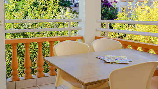 Bilde av en balkong med et bord og stoler på flisebelagt gulv, samt utsikt over vegetasjon og bygninger. (AI-generert tekst)