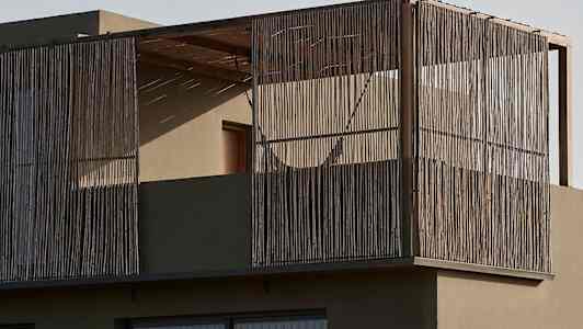 En moderne bygning med en balkong innkapslet med trestaver og glassvinduer. Foran ses vegetasjon i et ørkenlandskap. (AI-generert tekst)