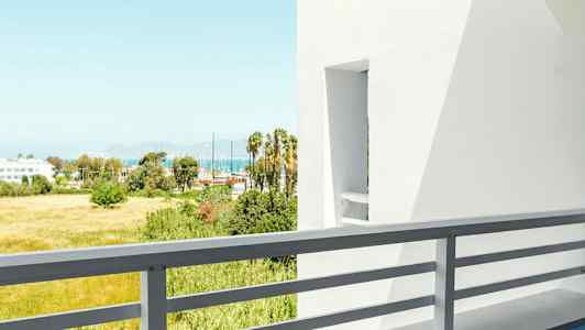 Utsikt fra en balkong med bord og stol, hvite bygninger og grøntområder i bakgrunnen. (AI-generert tekst)