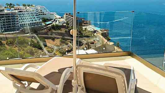 To solstoler under en parasoll på en terrasse med utsikt over havet og nærliggende hotell. (AI-generert tekst)