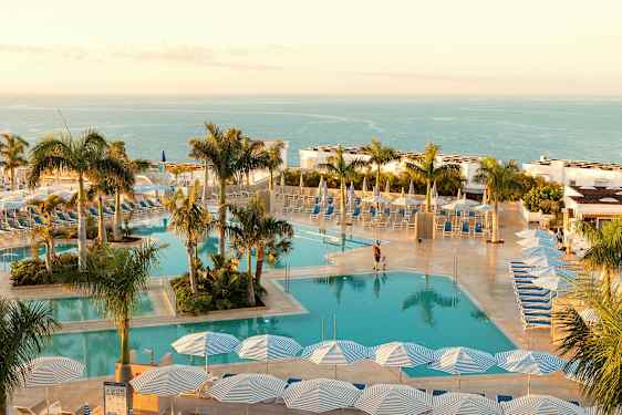 Et basseng med solsenger og parasoller, omgitt av palmer med havet i bakgrunnen. (AI-generert tekst)