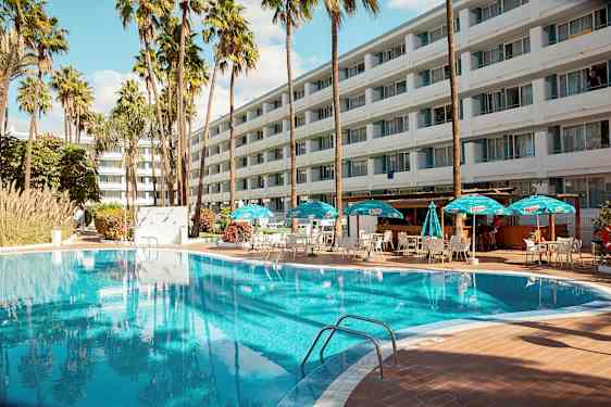 Utendørs svømmebasseng med solsenger og parasoller foran et stort hotell. (AI-generert tekst)