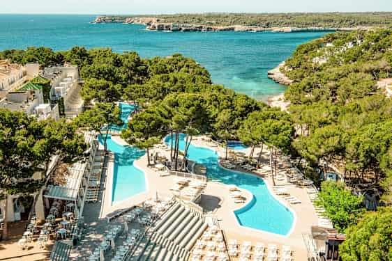 En utsikt over et feriested med bassenger omgitt av solsenger og parasoller, plassert ved siden av et turkist hav med frodig vegetasjon i bakgrunnen. (AI-generert tekst)