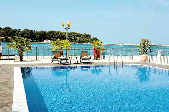 Et basseng med klart blått vann og to solsenger ved siden av. Havet er synlig i bakgrunnen. (AI-generert tekst)