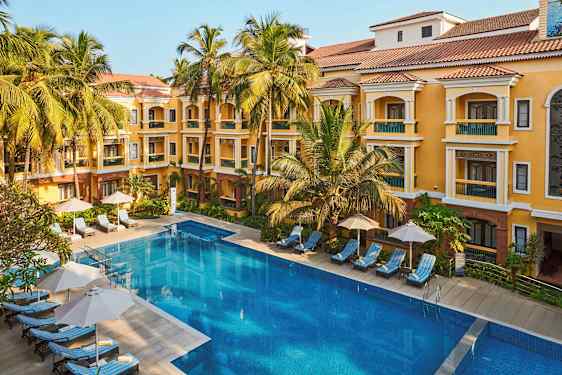 Et bilde av en hotellbygning med gul fasade og røde tak, omgitt av palmer. Foran bygningen er det et stort basseng og solsenger med parasoller. (AI-generert tekst)
