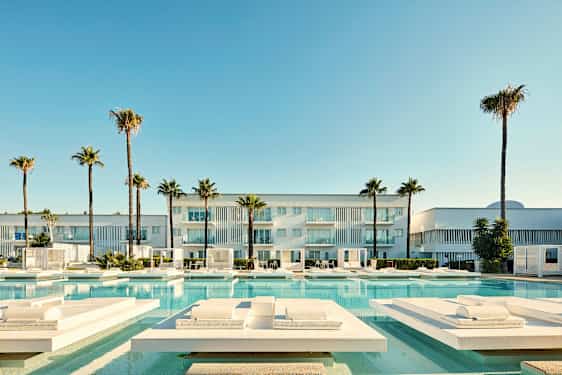 En moderne bygning med balkonger, omgitt av palmer og et svømmebasseng med solsenger. (AI-generert tekst)