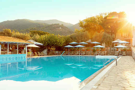 Et utendørs basseng med solsenger og parasoller, omgitt av trær og bygninger i et fjellandskap. (AI-generert tekst)