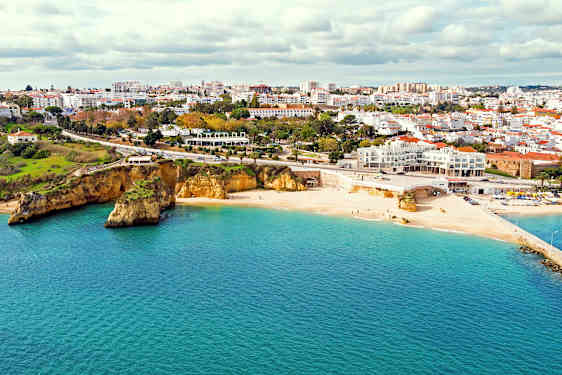 Lys sandstrand ved havet med en by i bakgrunnen og klipper ved stranden. (AI-generert tekst)