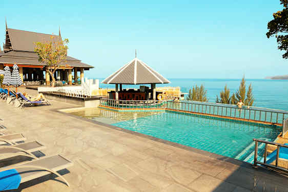 Et utendørs basseng med solsenger, parasoller og en bar ved siden. Utsikt over havet og trær i bakgrunnen. (AI-generert tekst)