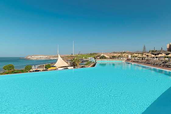 Et stort utendørsbasseng med solsenger og parasoller ved siden av, med utsikt over havet og en kystby. (AI-generert tekst)