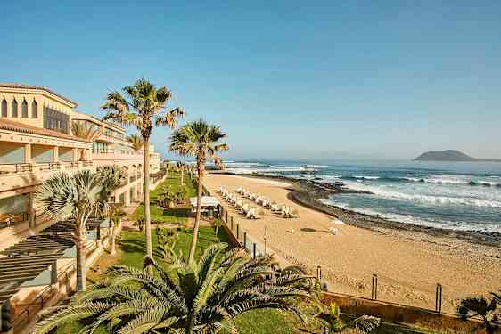 En strand med sand, parasoller og bølger, omgitt av bygninger og palmer. (AI-generert tekst)