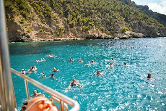 Svømmende mennesker i klart blått vann foran en klipperik kystlinje. (AI-generert tekst)