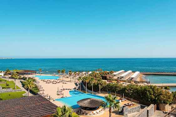 Utsikt over en resort med basseng, solsenger, palmer og hav i bakgrunnen. (AI-generert tekst)