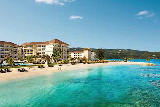 På stranden ligger et hotell med flere etasjer og palmetrær, ved siden av turkist hav og solsenger. (AI-generert tekst)
