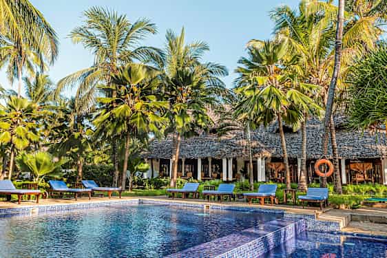 Et basseng omgitt av solsenger og palmer ved en bygning med stråtak. (AI-generert tekst)