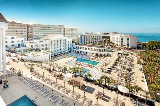 Utsikt over et hotellområde med bassenger, solsenger og hvit arkitektur nær havet. (AI-generert tekst)