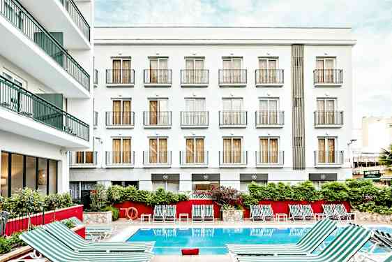 Bilde av en hotellbygning med hvit fasade og flere balkonger. Foran bygningen er et utendørsbasseng med stripete solsenger plassert rundt. (AI-generert tekst)