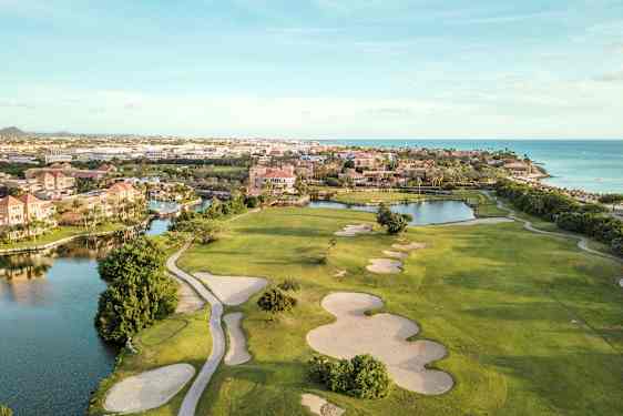 Golfbane med sandbunkere og grønne plenområder nær vannløp og strand. Bygninger i bakgrunnen. (AI-generert tekst)